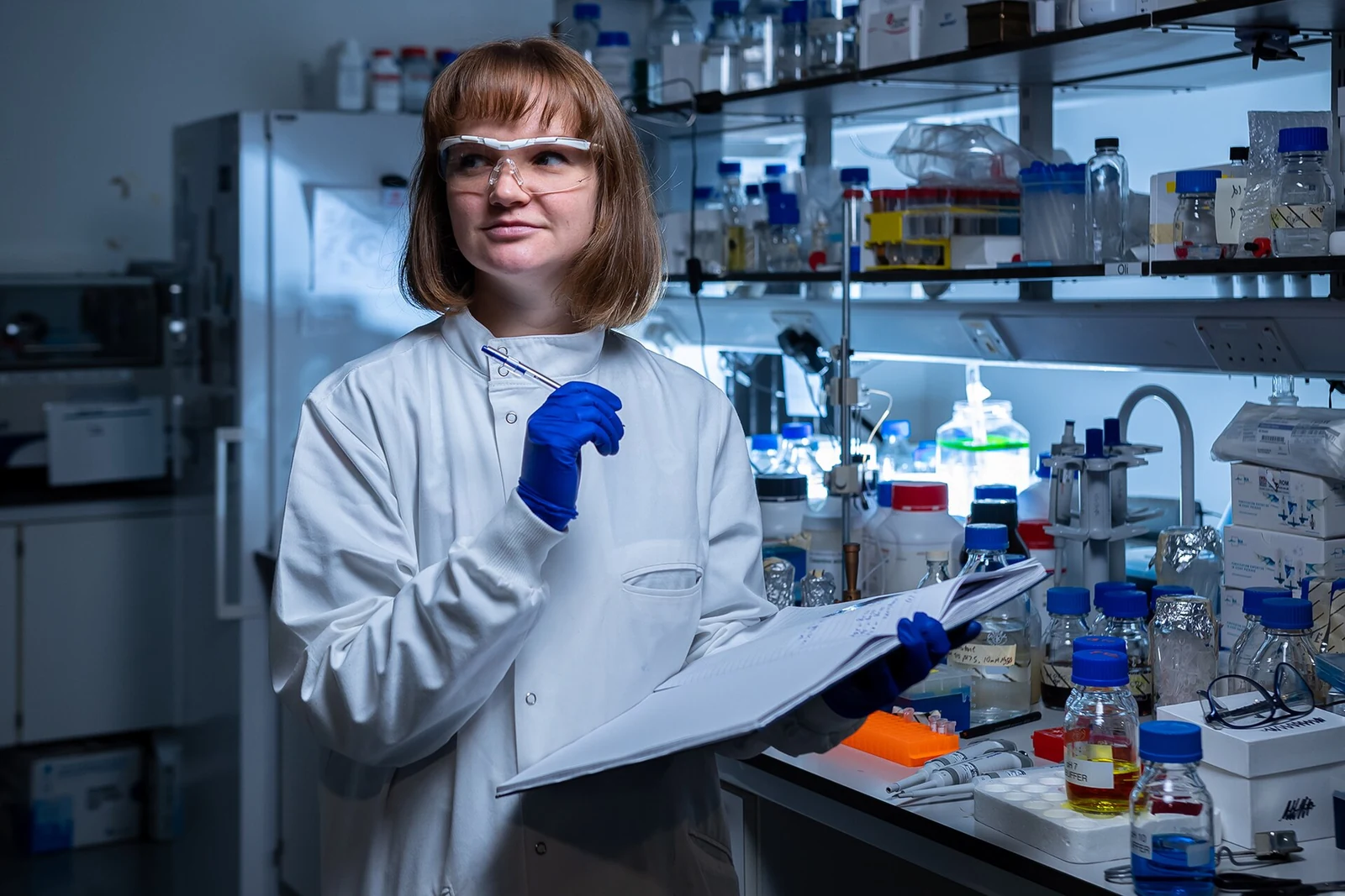 A researcher in safety glasses and blue gloves holds an open notebook and pen beside a laboratory bench.