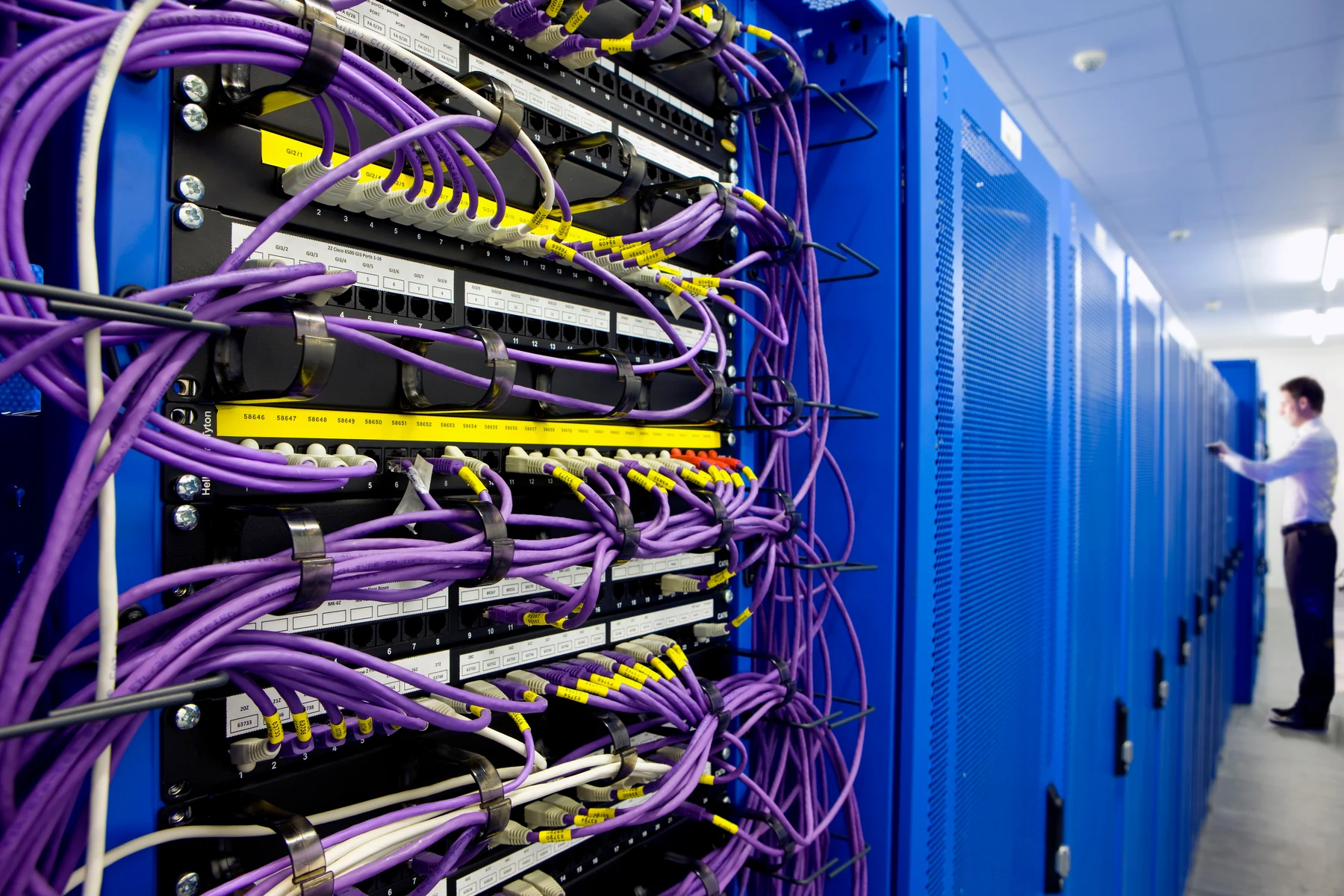 Purple network patch cables connected to labelled rack ports, with blue cabinets and a person farther along the aisle.