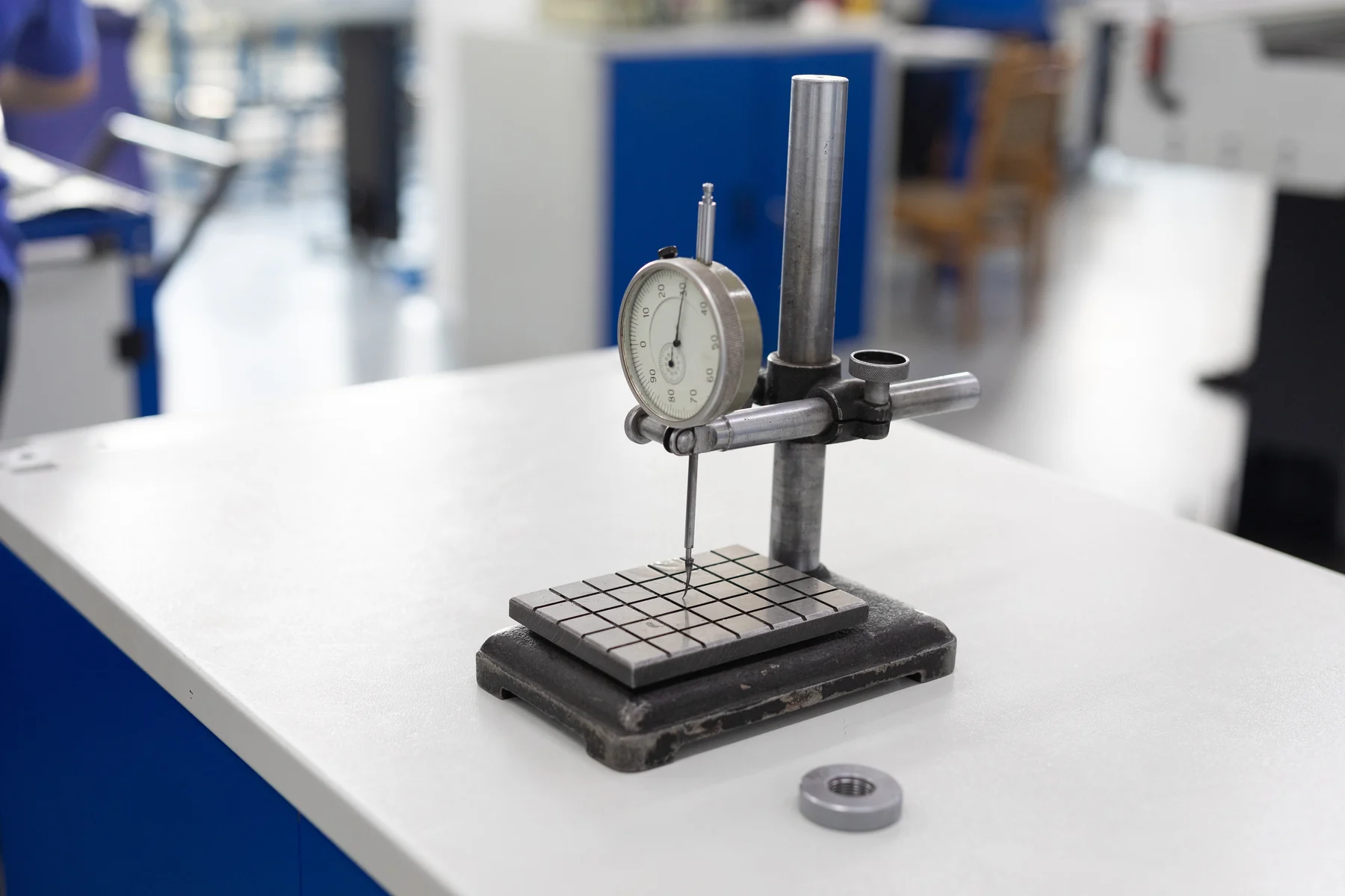 An analogue dial indicator on a metal stand above a grooved base in a workshop.