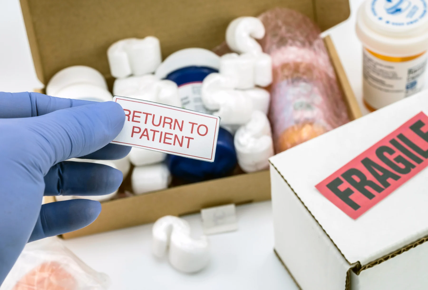 A blue-gloved hand holding a Return to patient label beside a carton of medicine-style containers.