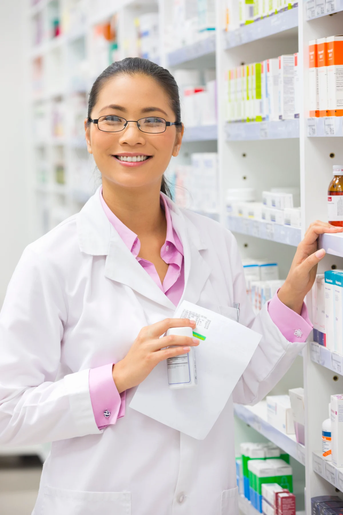 A person in a white coat holding a white bottle and paper beside pharmacy-style shelves.