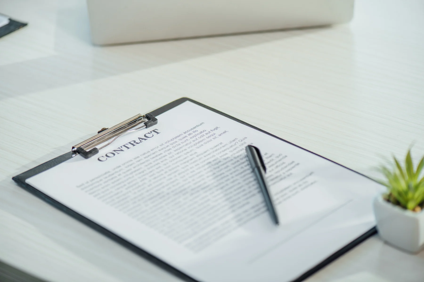 A black clipboard with a paper headed Contract and a pen on a pale wooden table.
