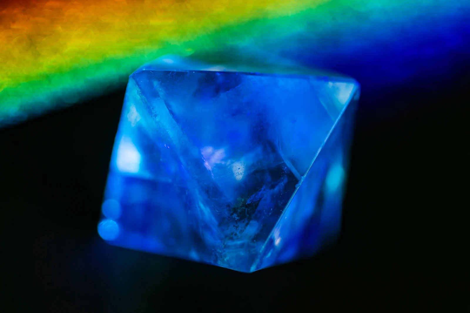A blue-lit faceted transparent crystal sits below a band of rainbow-coloured light against black.